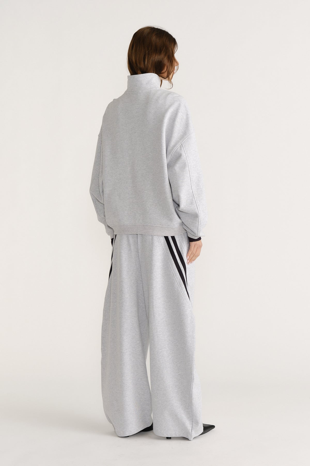 Woman seen from behind wearing a relaxed light grey quarter zip sweat with matching wide-leg pants featuring black side stripes