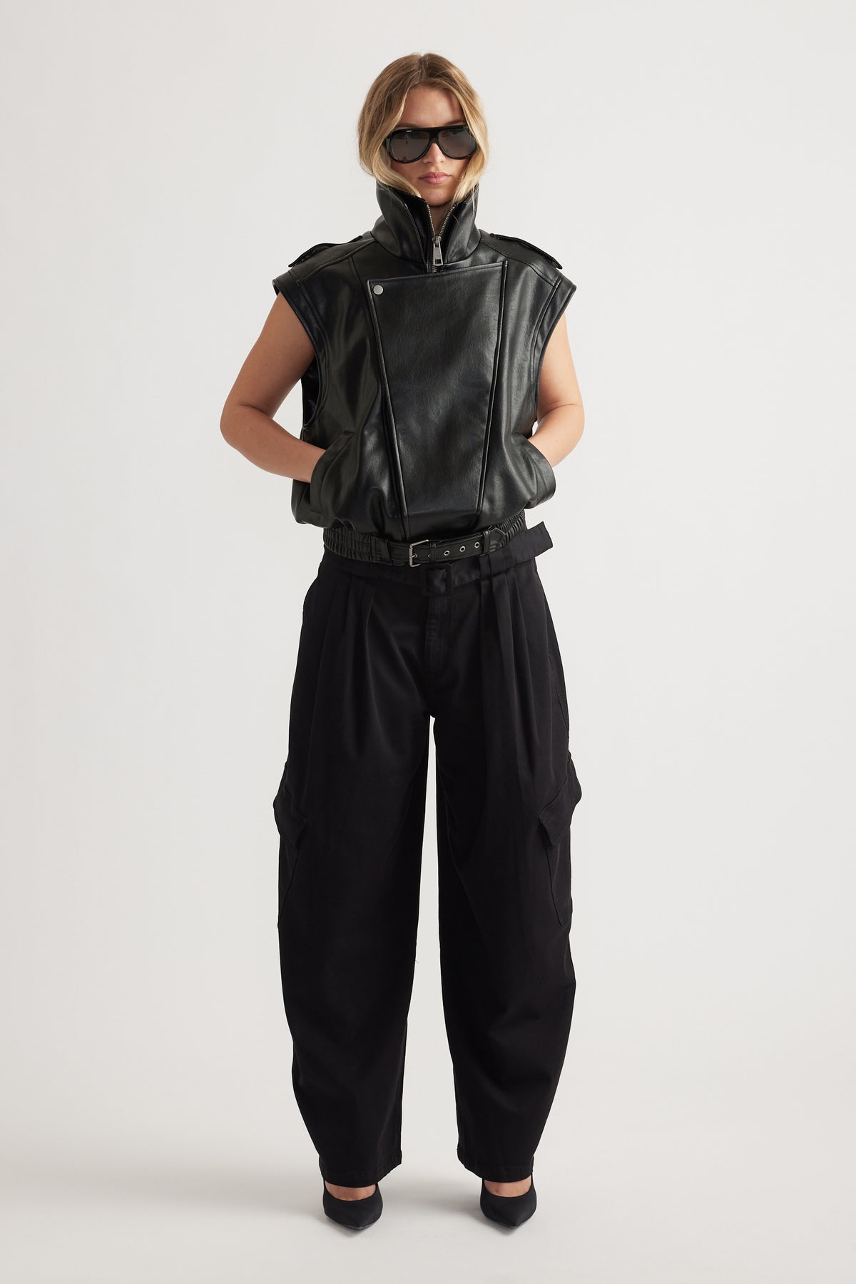 Woman styled in sleeveless black leather jacket with high collar and loose black balloon cargo pants with side pockets