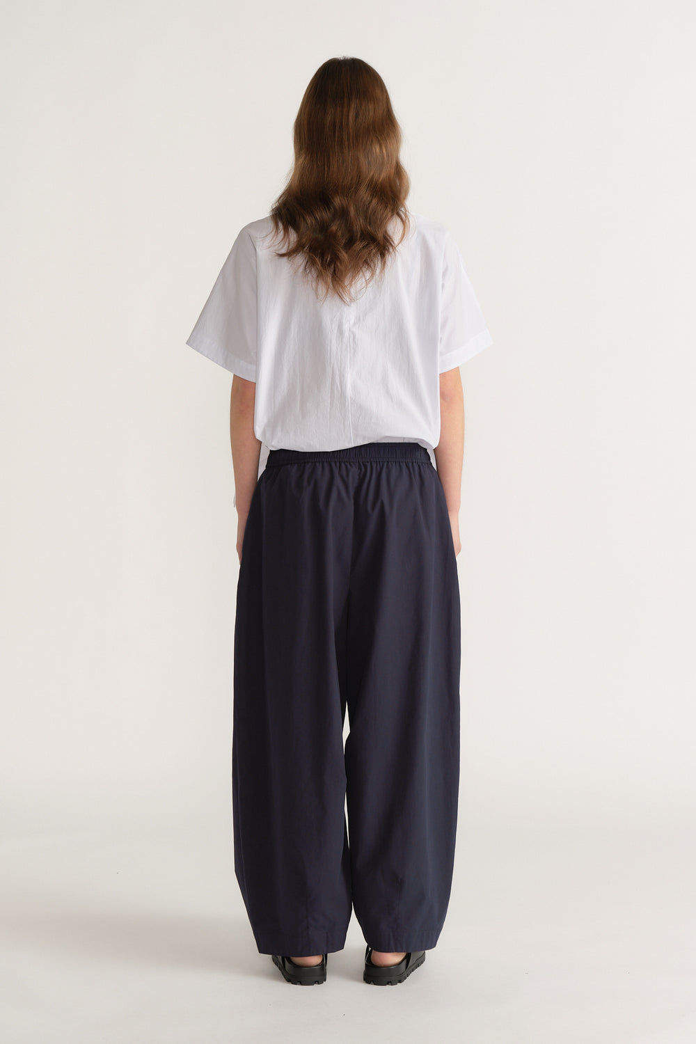 Woman wearing a white short sleeve top with navy barrel leg pants and black shoes, viewed from behind