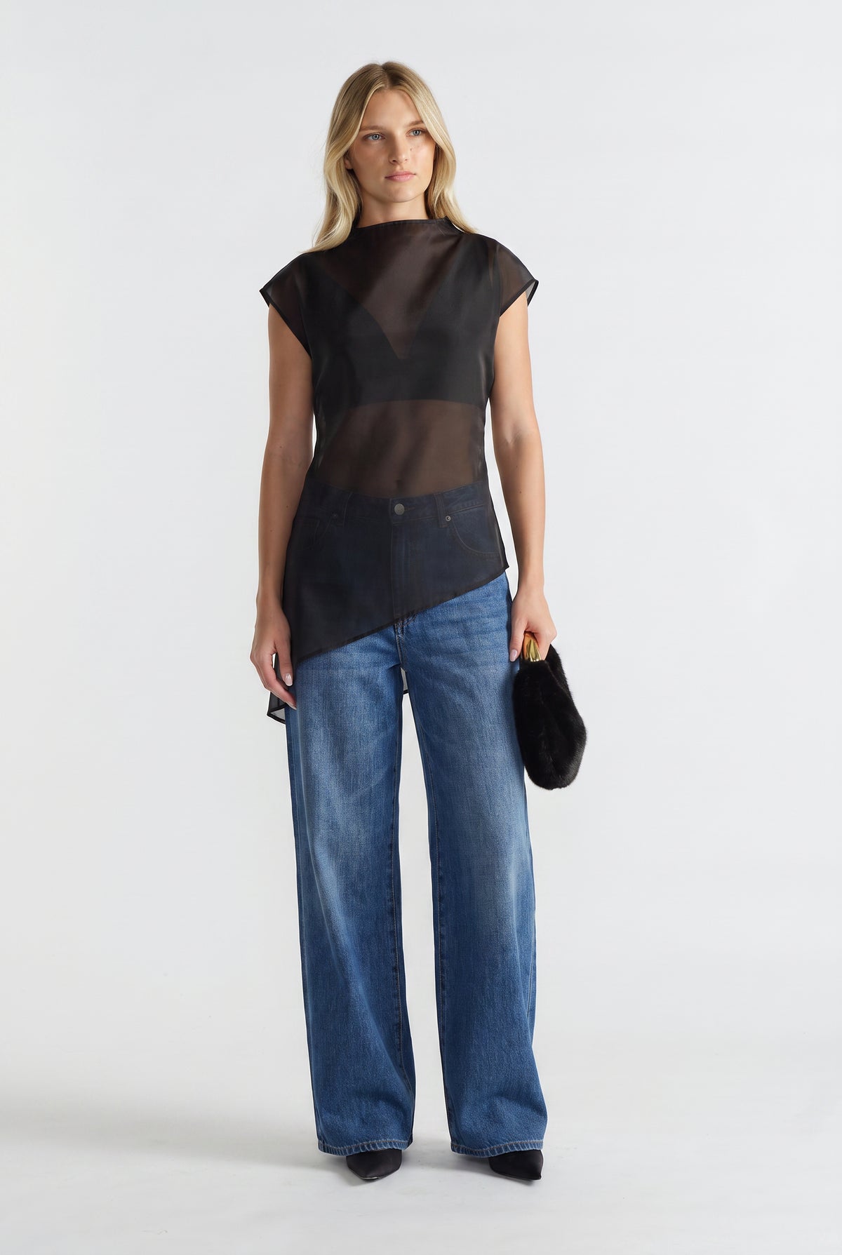 Woman wearing a black sheer high neck sleeveless tie waist top with an asymmetric hem, wide leg jeans and a black handbag