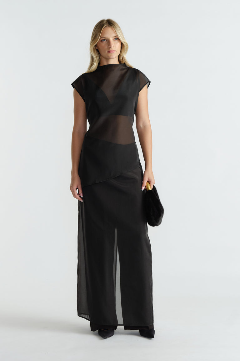 Woman wearing a black sheer sleeveless top with a high neckline and asymmetric hem, styled with wide leg pants