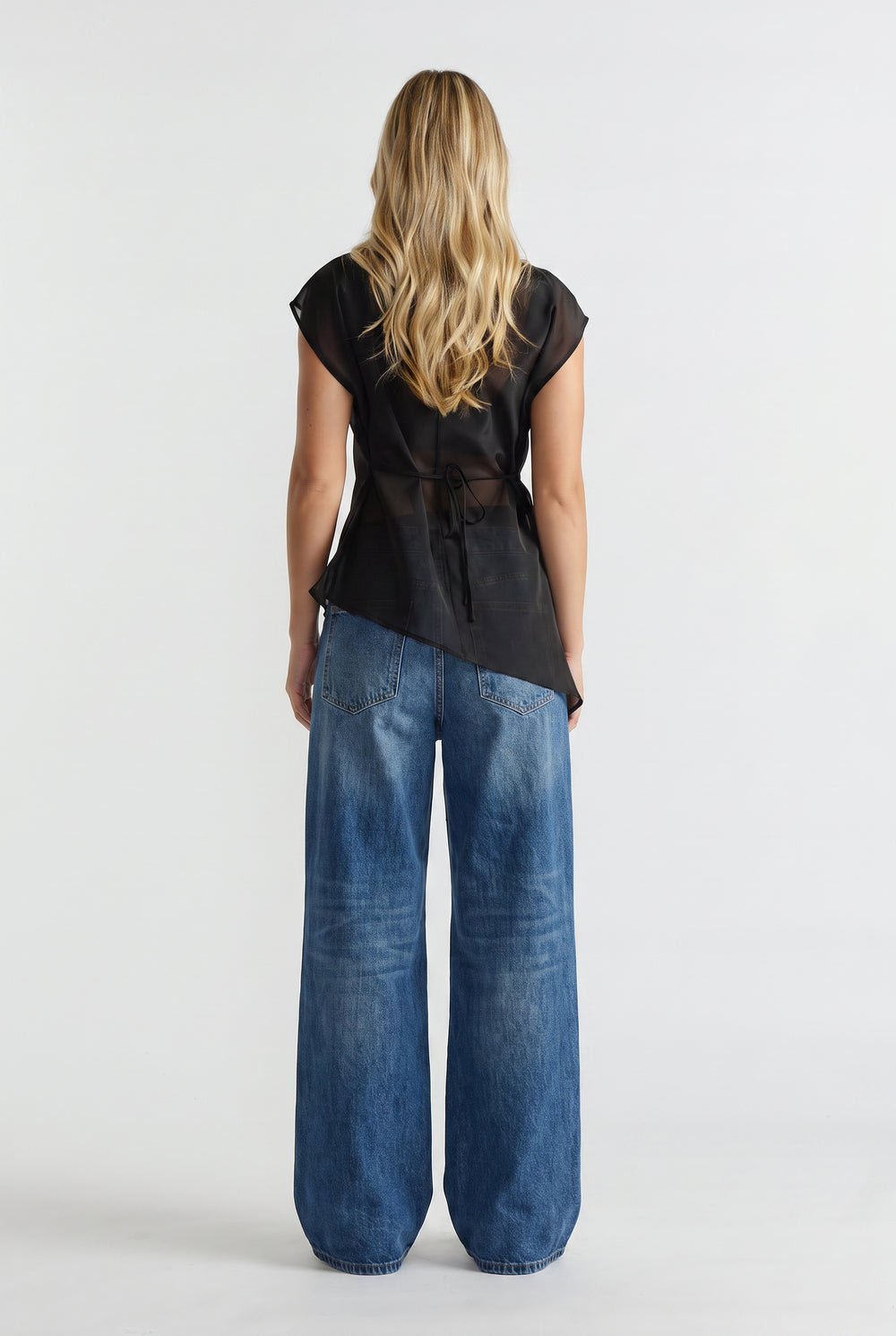 Woman wearing a black sheer tie waist top with an asymmetric hem and wide leg blue jeans