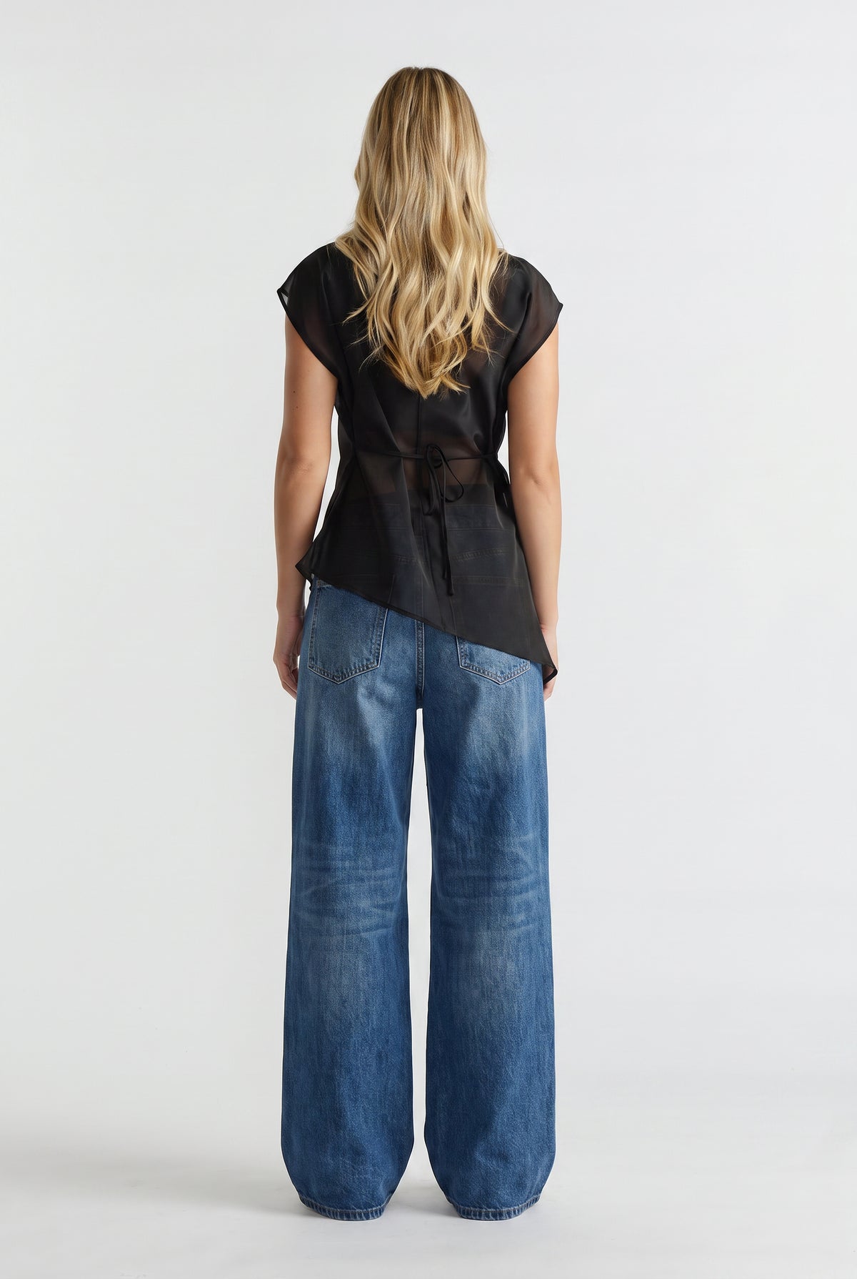 Woman wearing a black sheer tie waist top with an asymmetric hem and wide leg blue jeans