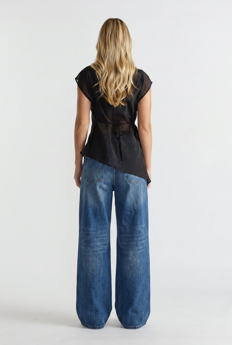 Woman wearing a black sheer tie waist top with an asymmetric hem and wide leg blue jeans