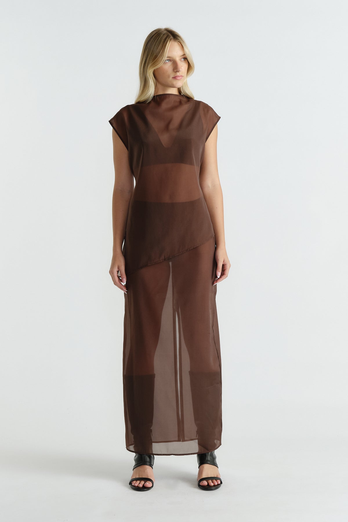Woman wearing a sleeveless brown sheer dress with an asymmetric hem and black open-toe heeled sandals