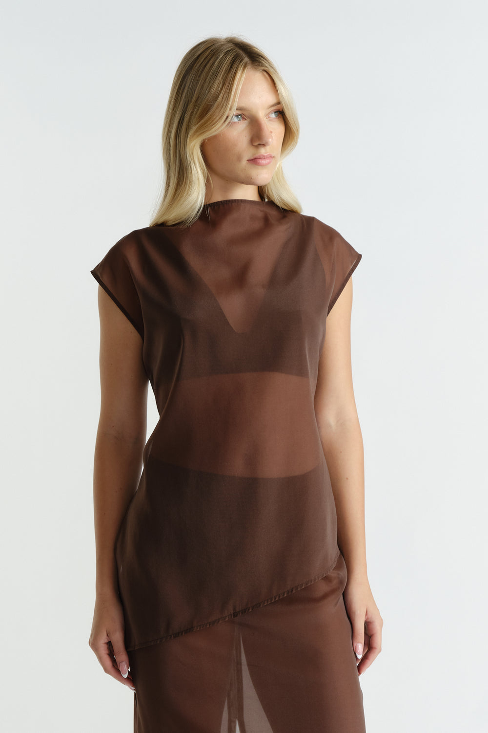 Woman wearing a sleeveless brown sheer top with an asymmetric hem and high neckline, styled with a bralette underneath