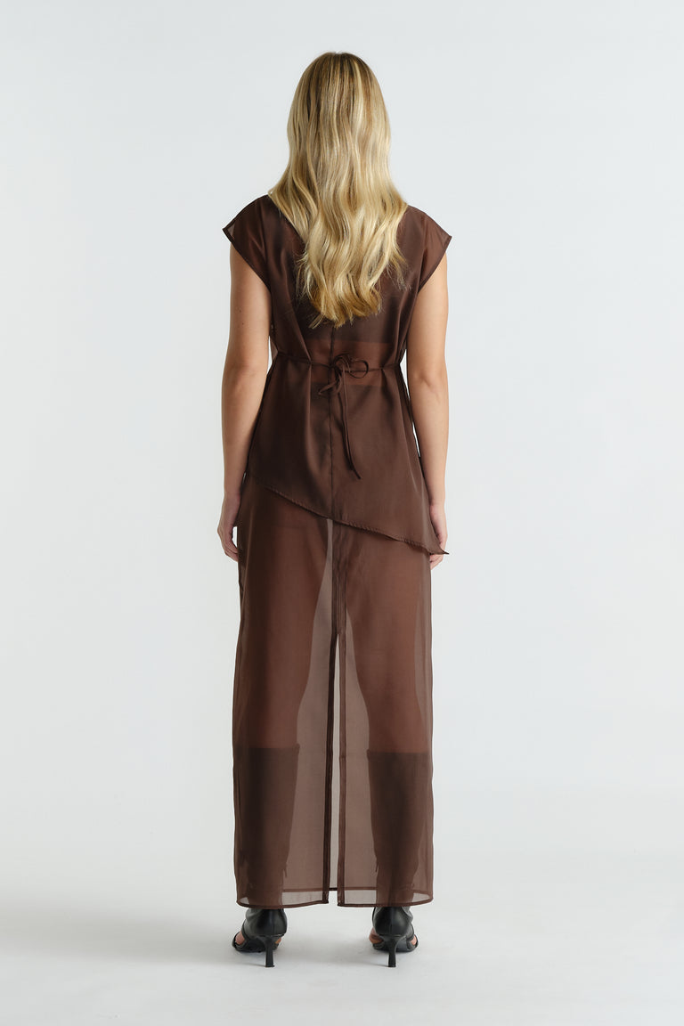 Rear view of woman wearing sleeveless brown sheer dress with waist tie and asymmetric hem paired with black heeled sandals