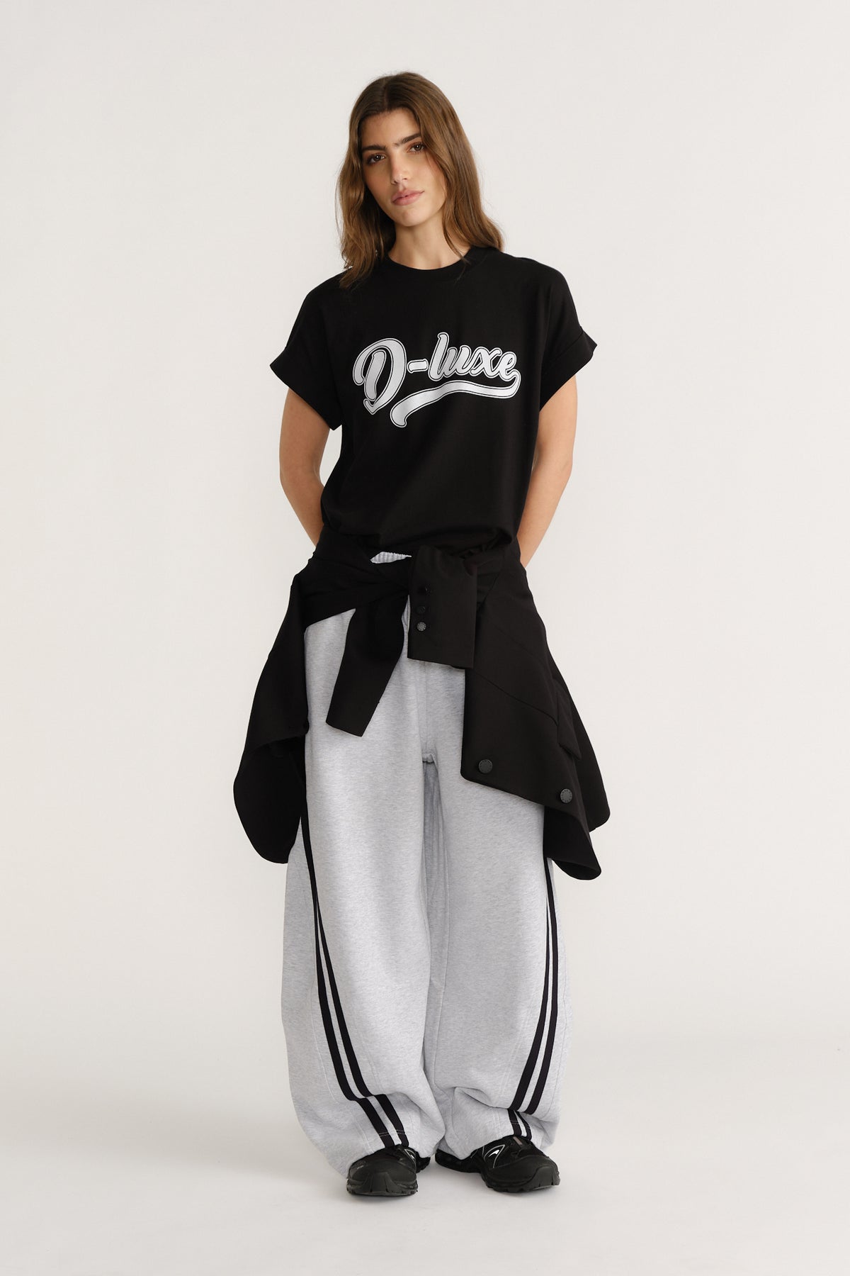 Woman wearing a relaxed black graphic tee paired with loose grey track pants featuring side stripes and a black jacket tied at the waist