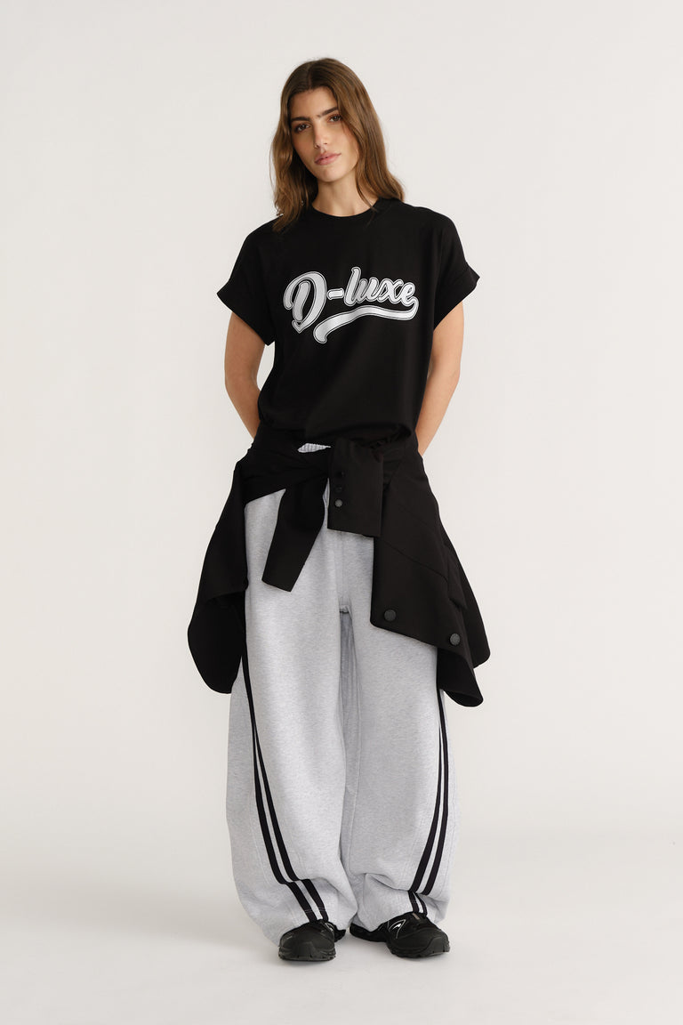 Woman wearing a relaxed black graphic tee paired with loose grey track pants featuring side stripes and a black jacket tied at the waist