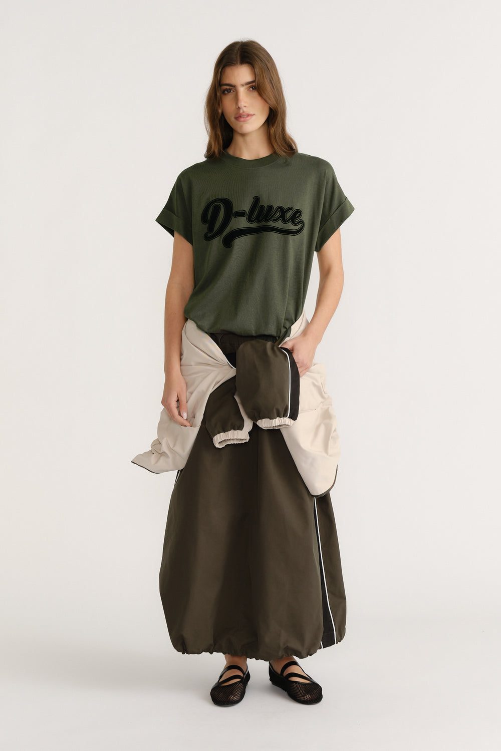 Woman wearing an olive green D-Luxe logo tee paired with a long dark skirt and black ballet flats.