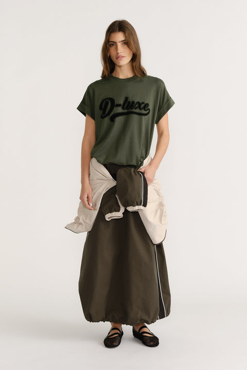 Woman wearing an olive green D-Luxe logo tee paired with a long dark skirt and black ballet flats.