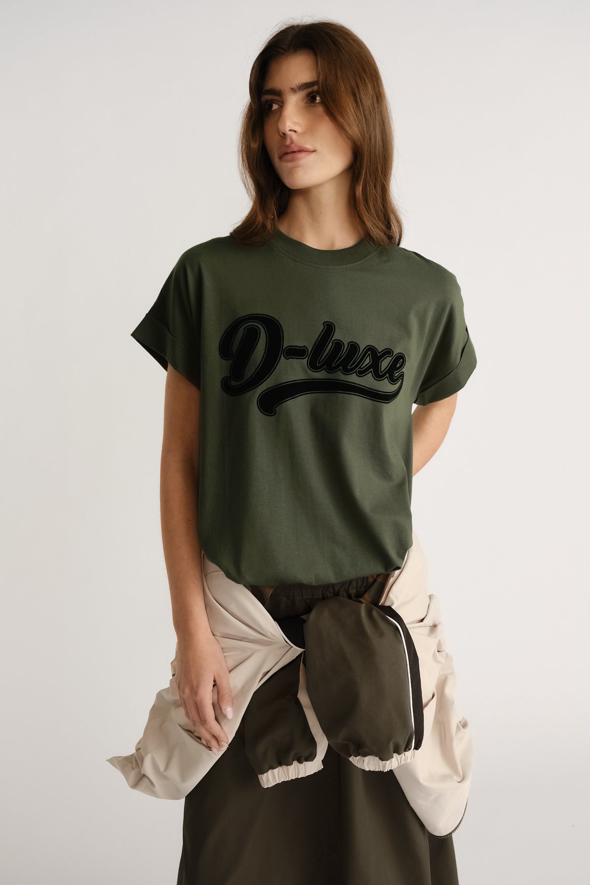 Woman wearing an olive green tee with bold graphic lettering, styled casually with a tied light jacket around the waist.
