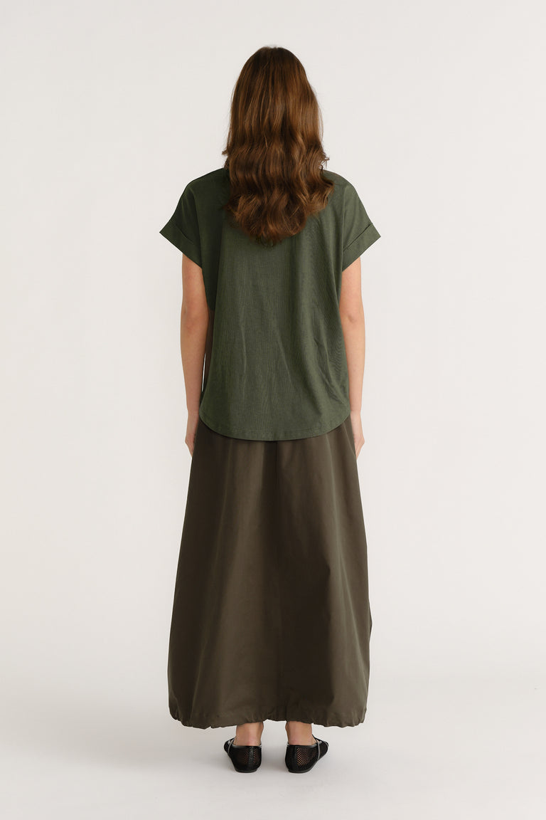 Woman wearing relaxed green tee paired with a flowing olive maxi skirt and black flat shoes, shown from behind.