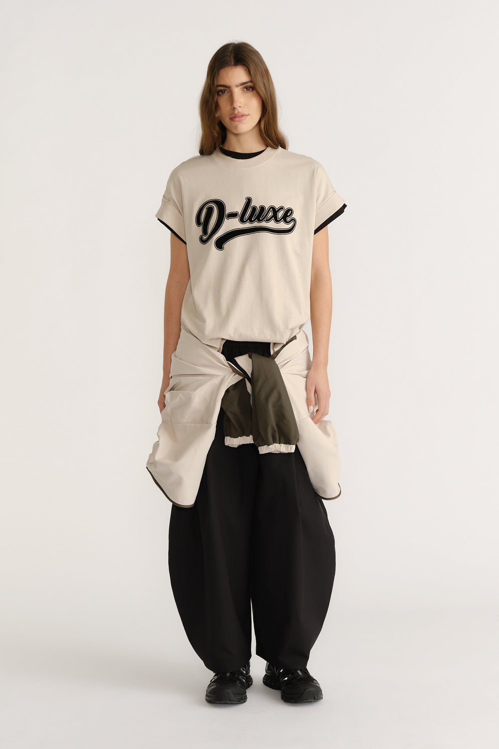 Woman wearing a relaxed beige tee with black trim and script logo, paired with wide black pants and a tied jacket.