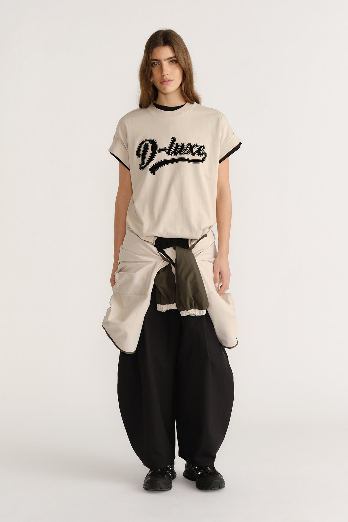Woman wearing a relaxed beige tee with black trim and script logo, paired with wide black pants and a tied jacket.