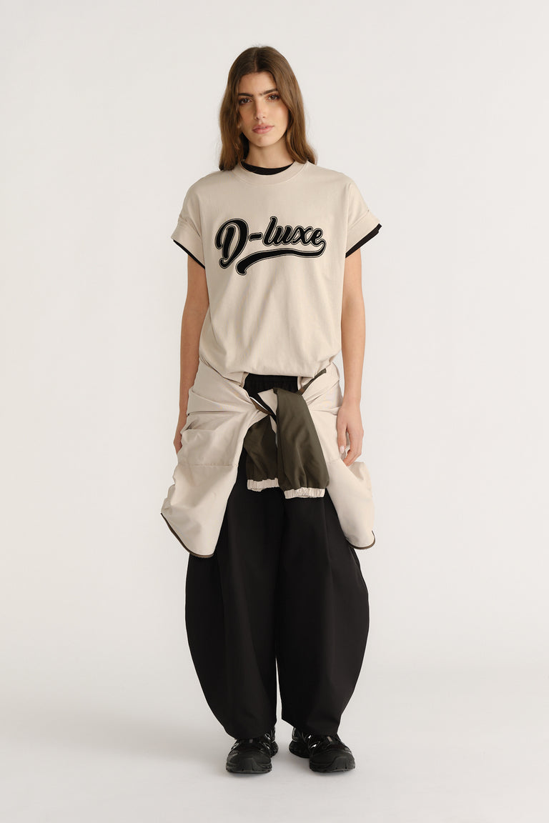 Woman wearing a relaxed beige tee with black trim and script logo, paired with wide black pants and a tied jacket.