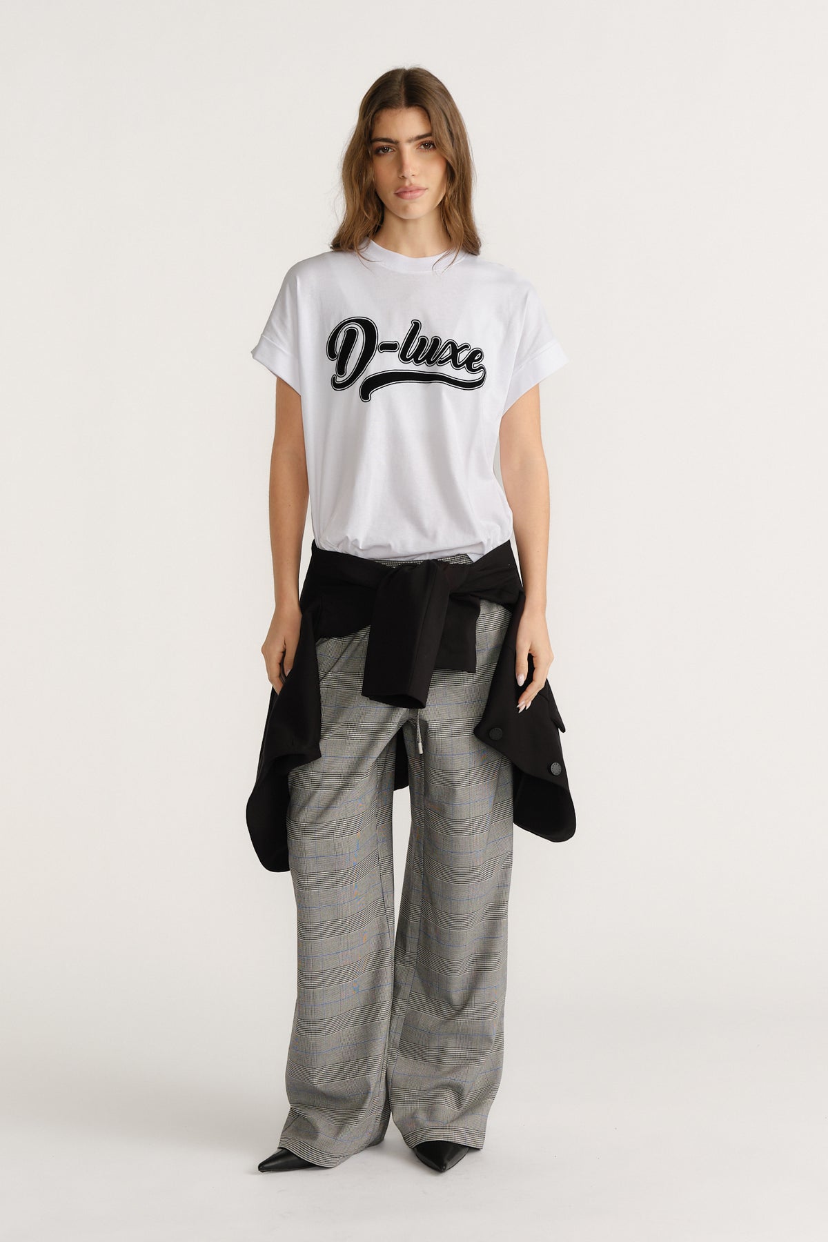 Woman wearing a white graphic tee paired with grey plaid wide-leg pants and a black jacket tied around the waist