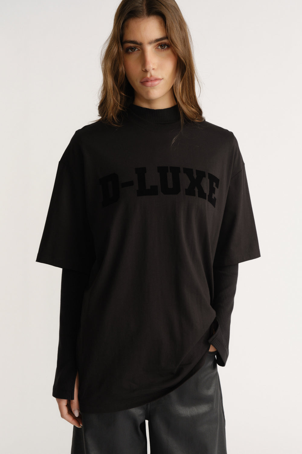 Woman wearing a relaxed black tee with flocked "D-LUXE" logo layered over a long-sleeve black top and leather pants.