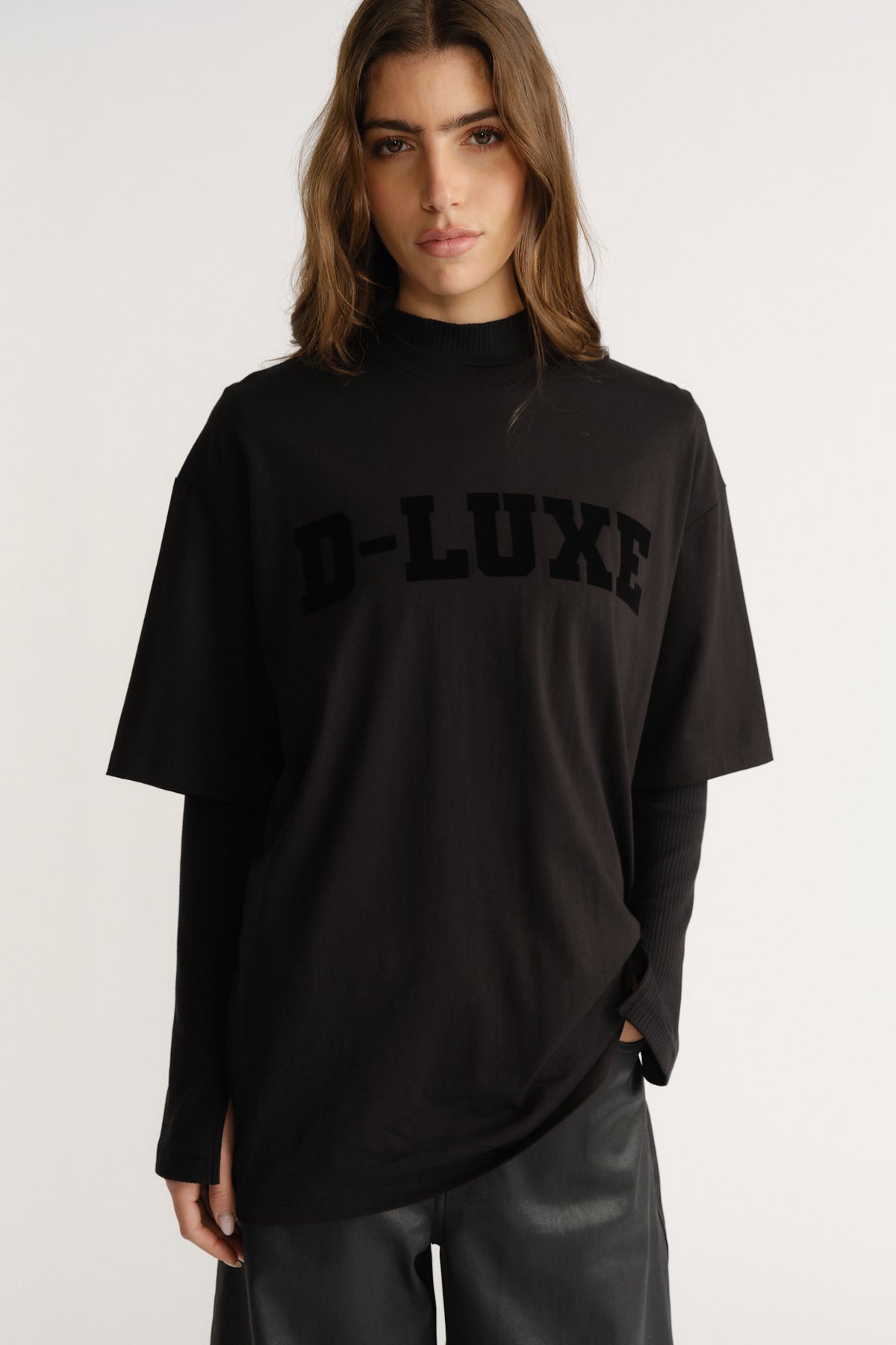 Woman wearing a relaxed black tee with flocked "D-LUXE" logo layered over a long-sleeve black top and leather pants.