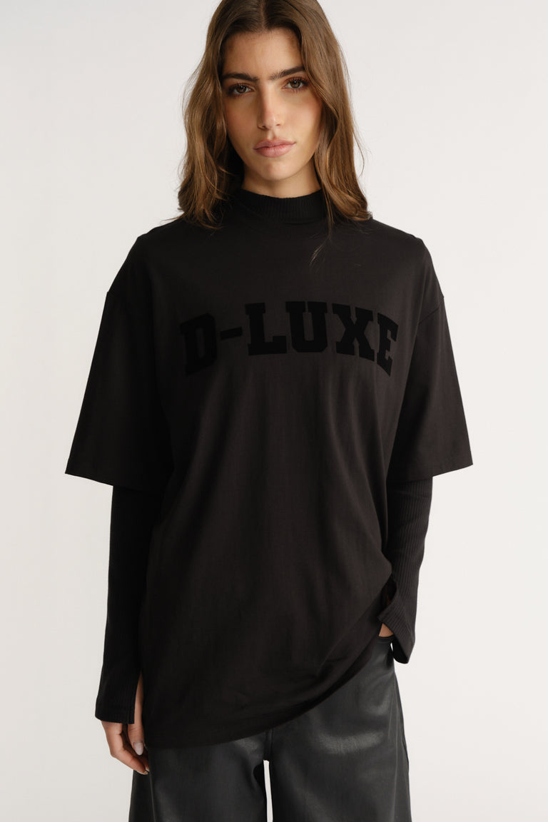 Woman wearing a relaxed black tee with flocked "D-LUXE" logo layered over a long-sleeve black top and leather pants.