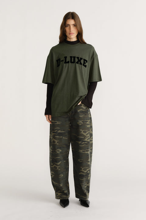 Woman wearing olive green tee with bold logo paired with loose-fitting camouflage pants and black long-sleeve layer underneath