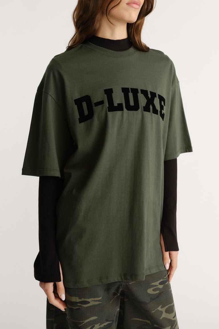Woman wearing an olive green oversized tee with bold black flocked lettering styled over a black long-sleeve top.
