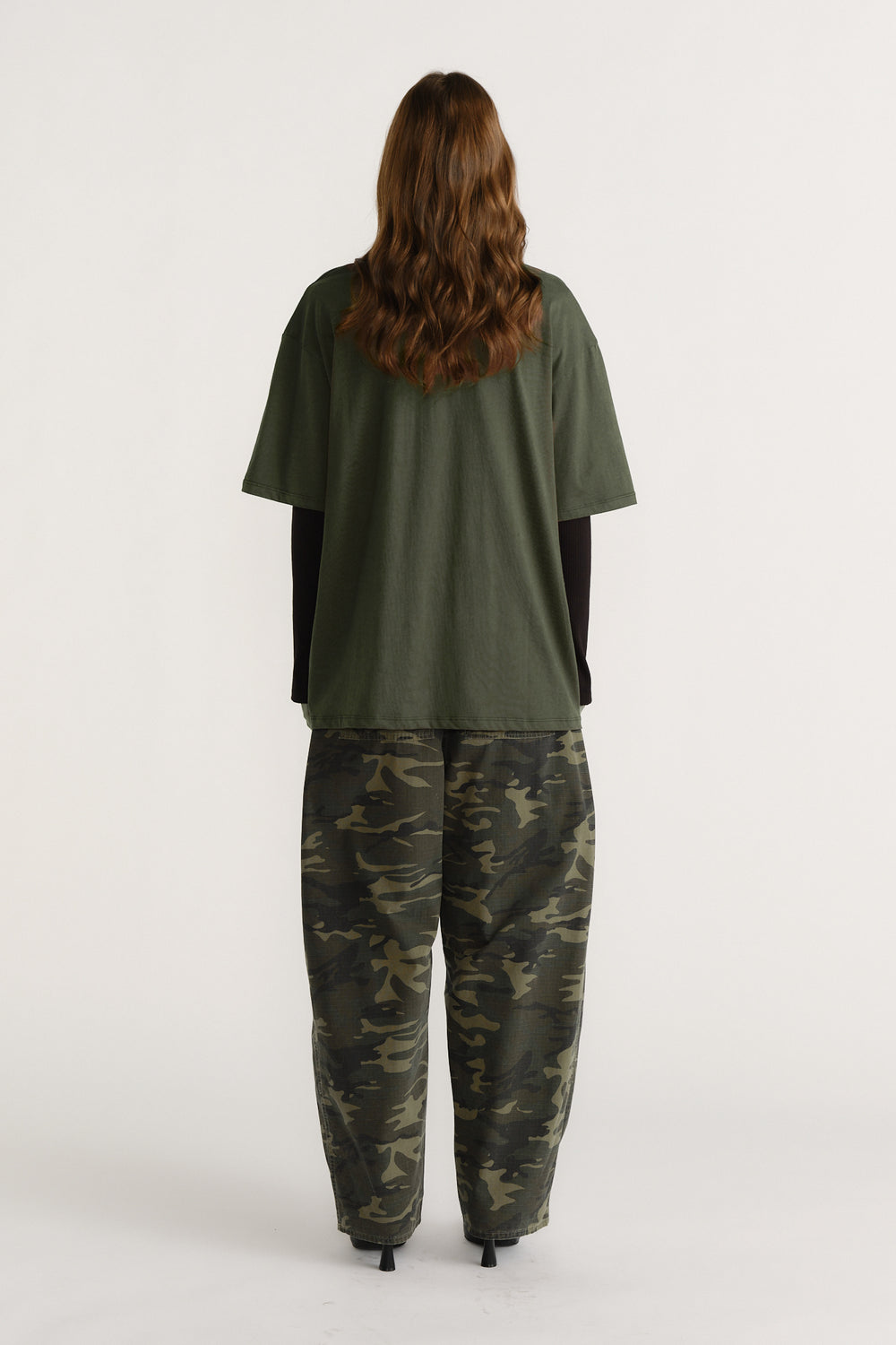Woman wearing an olive green tee layered over a black long sleeve top paired with relaxed camo print pants, facing away.
