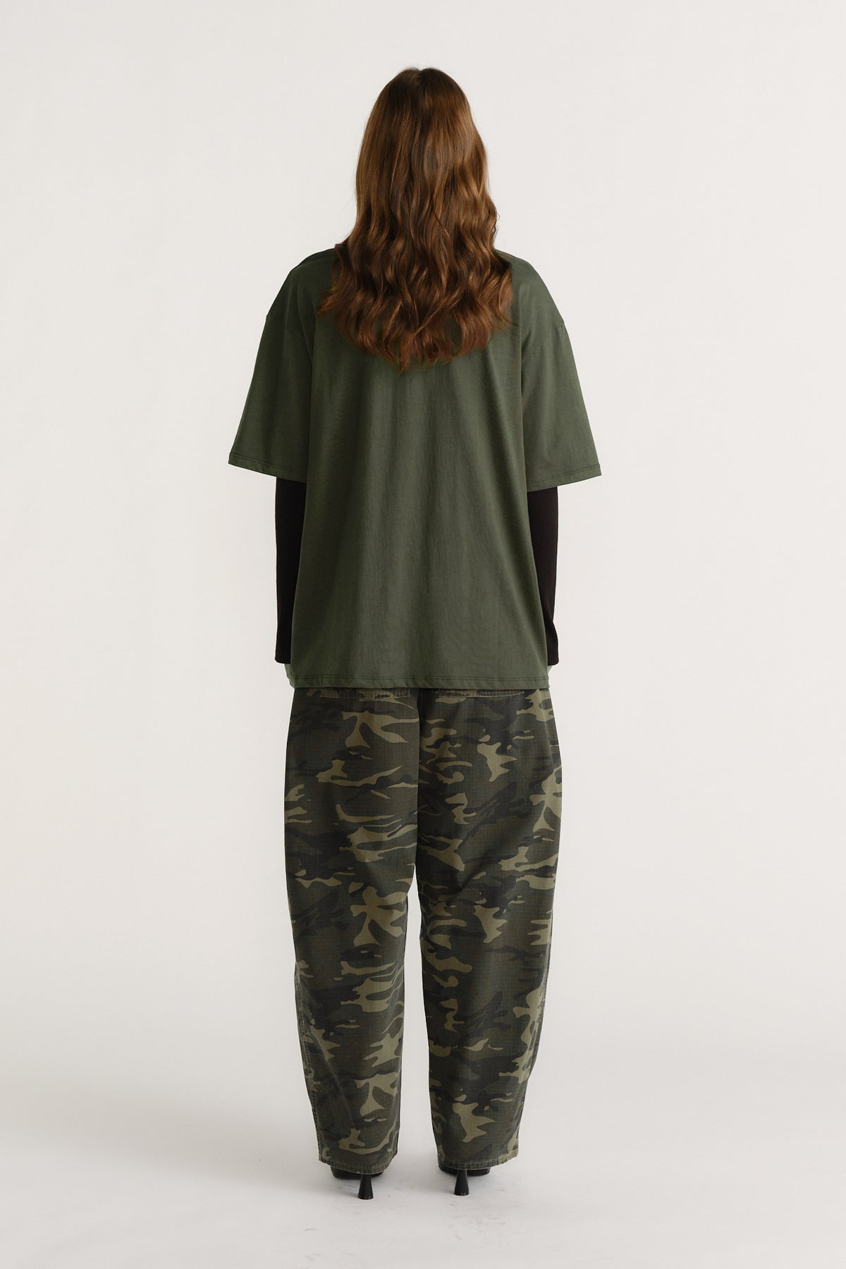 Woman wearing an olive green tee layered over a black long sleeve top paired with relaxed camo print pants, facing away.
