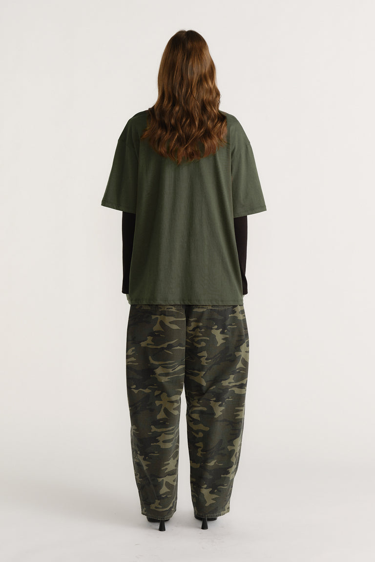 Woman wearing an olive green tee layered over a black long sleeve top paired with relaxed camo print pants, facing away.
