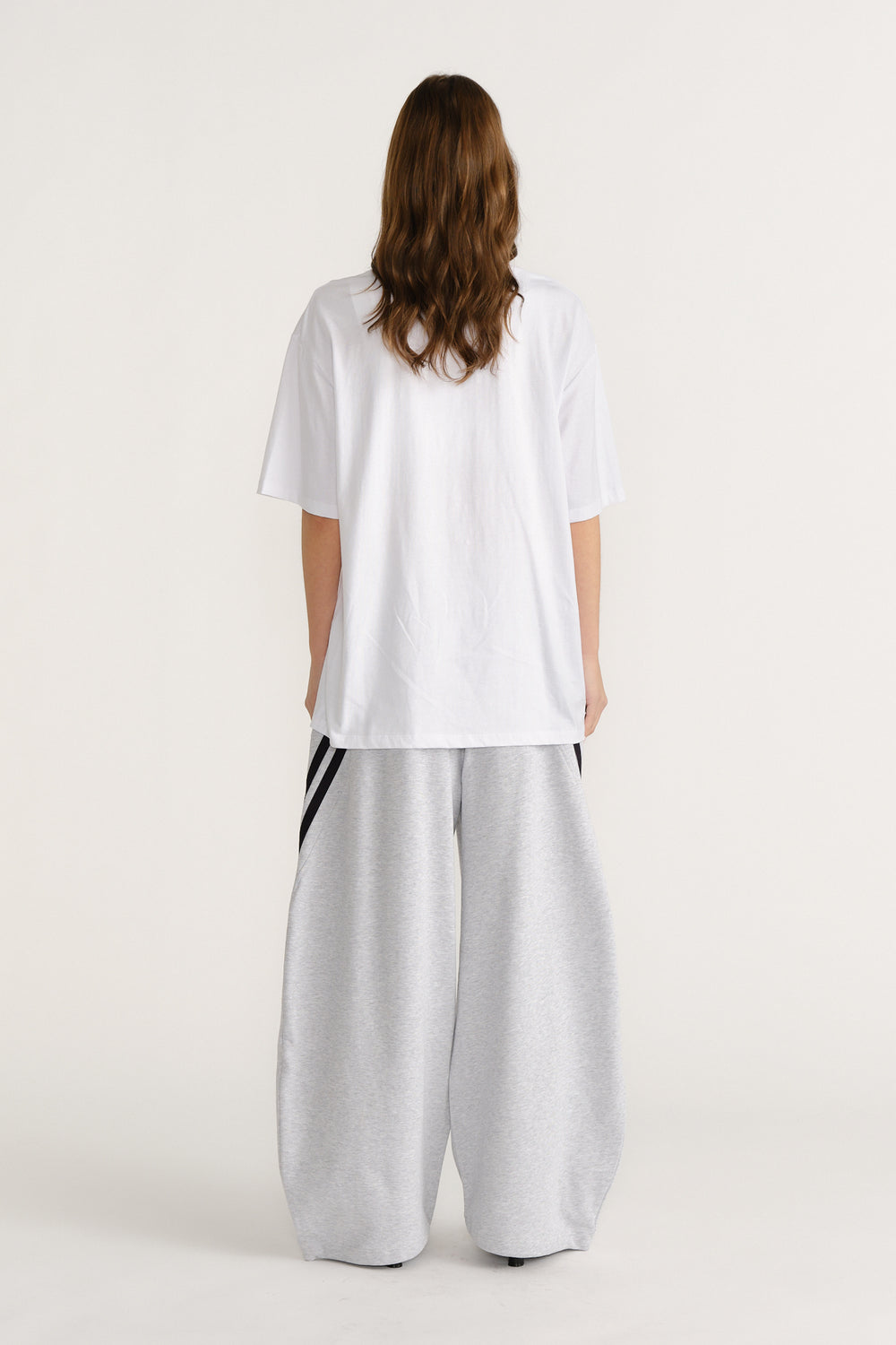 Back view of model wearing loose white tee paired with wide-leg grey pants featuring black side stripes.