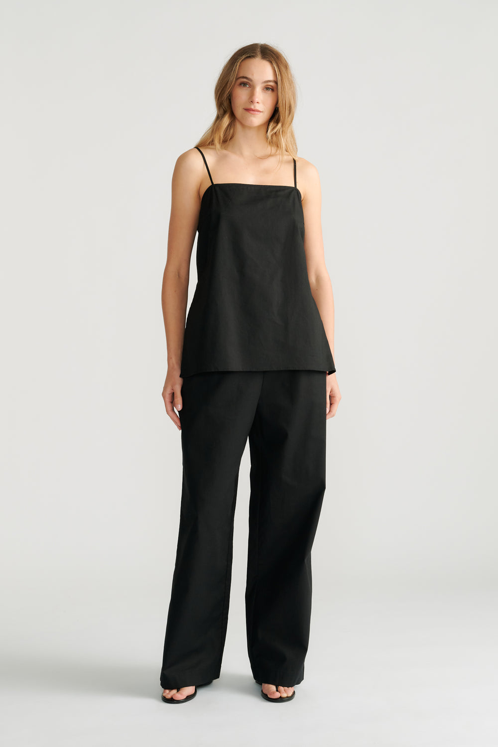 Woman wearing a sleek black camisole with thin straps paired with relaxed wide-leg black pants and black sandals.