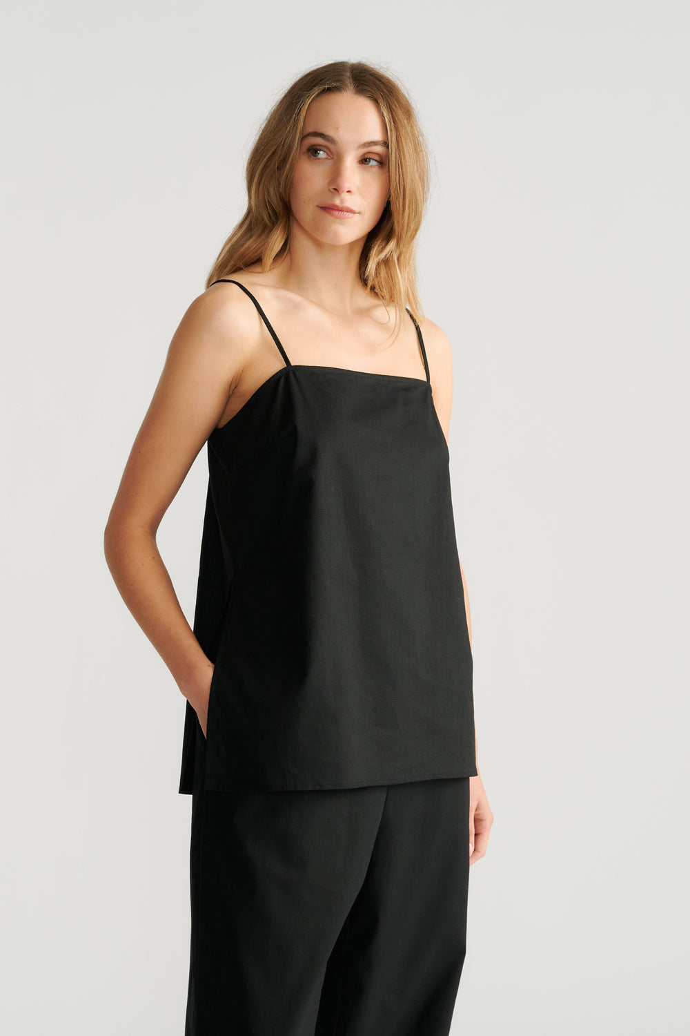 Woman wearing a relaxed black camisole with thin straps paired with loose black trousers, hands in pockets.