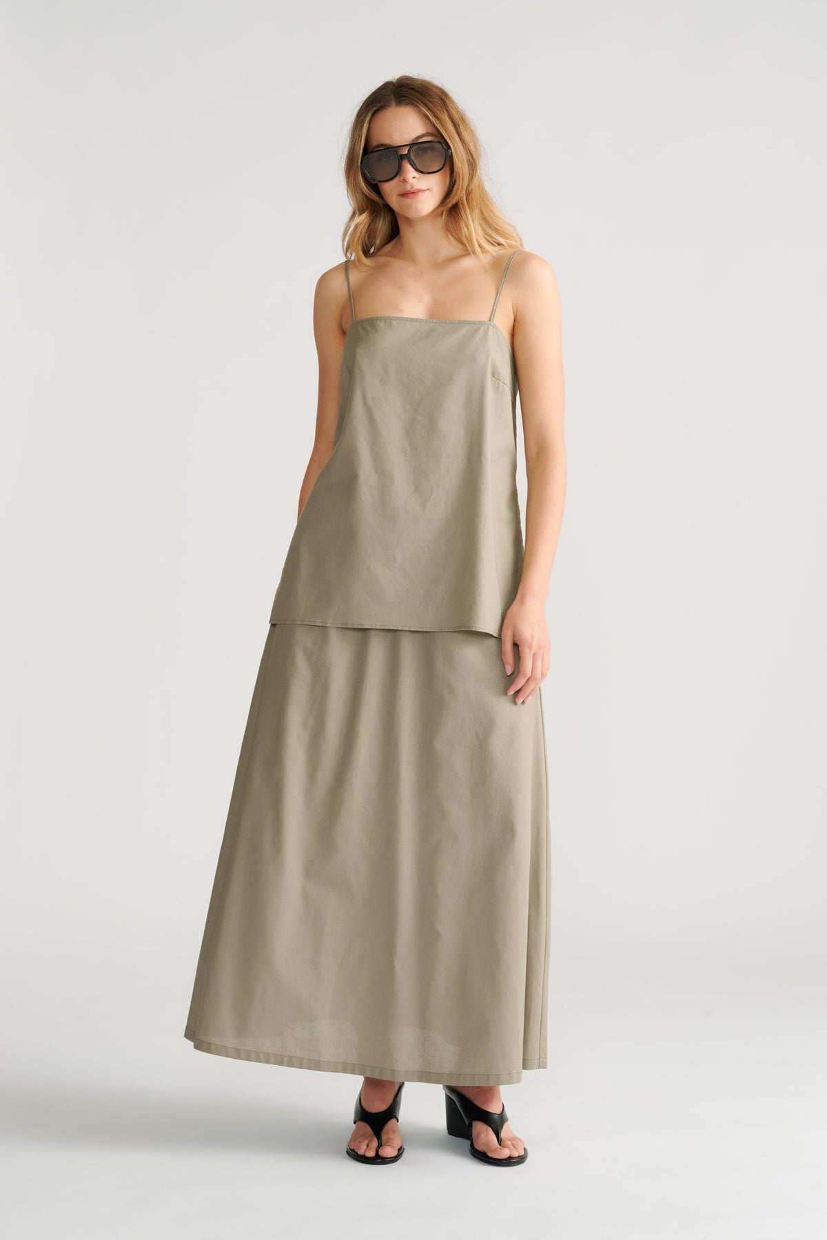 Woman wearing a sage green sleeveless cami layered over a matching long skirt with black heeled sandals and sunglasses.