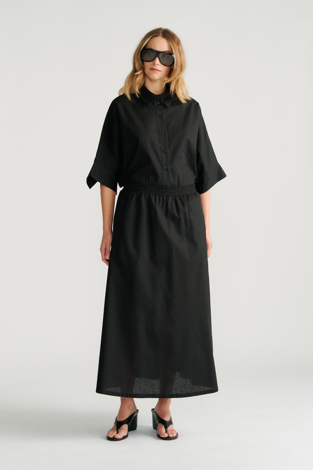 Woman wearing a black button-up top with elbow-length sleeves and a matching midi skirt paired with black heels and sunglasses.