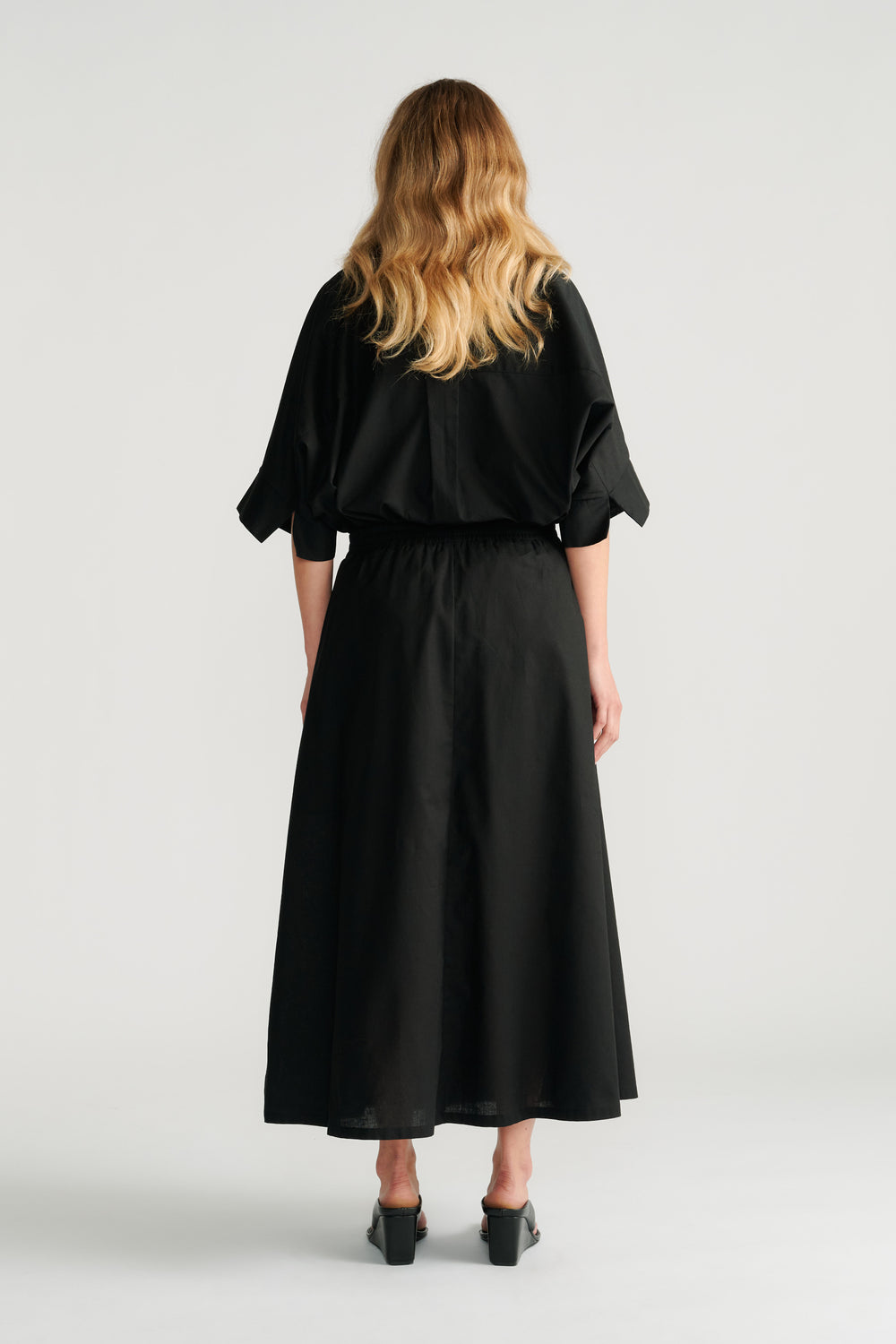 Woman wearing a relaxed black midi skirt with an elastic waistband paired with a loose black top and black heels, back view.