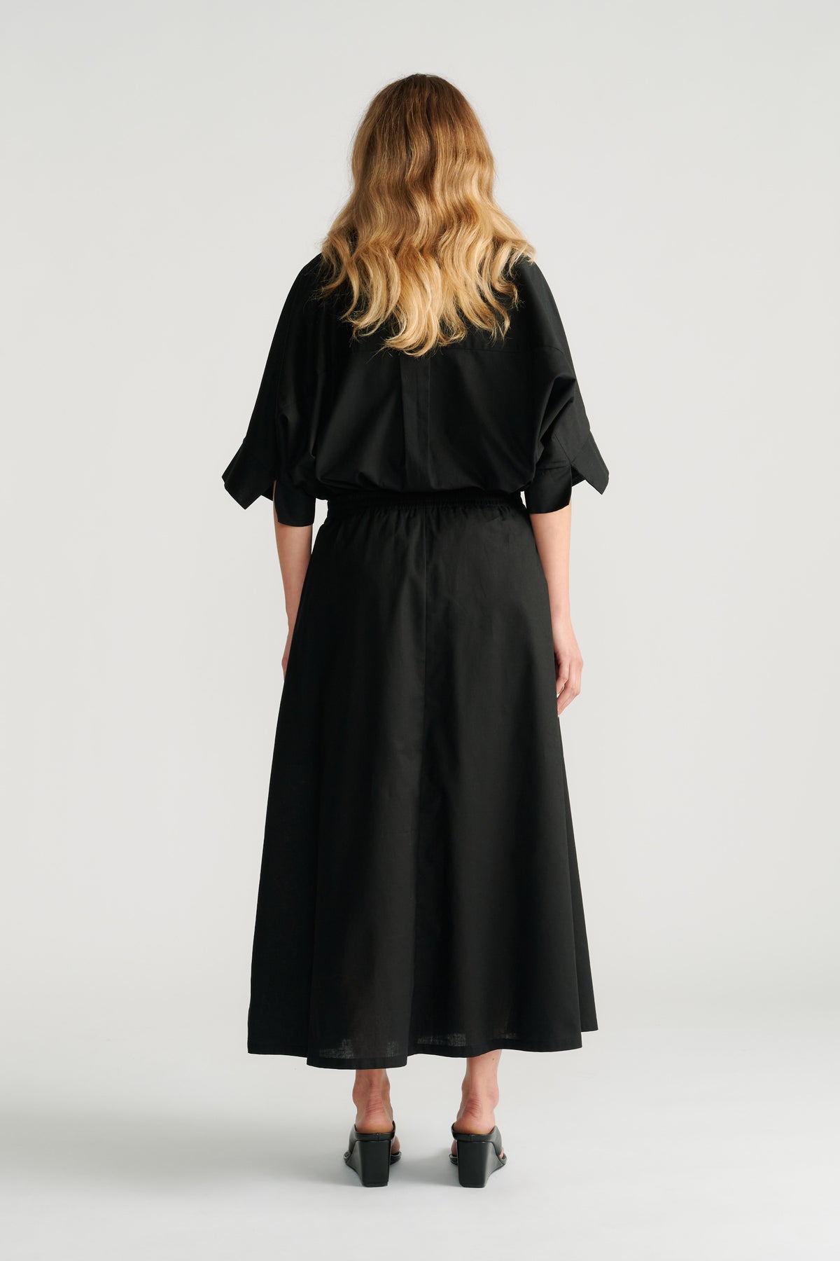 Woman wearing a relaxed black midi skirt with an elastic waistband paired with a loose black top and black heels, back view.