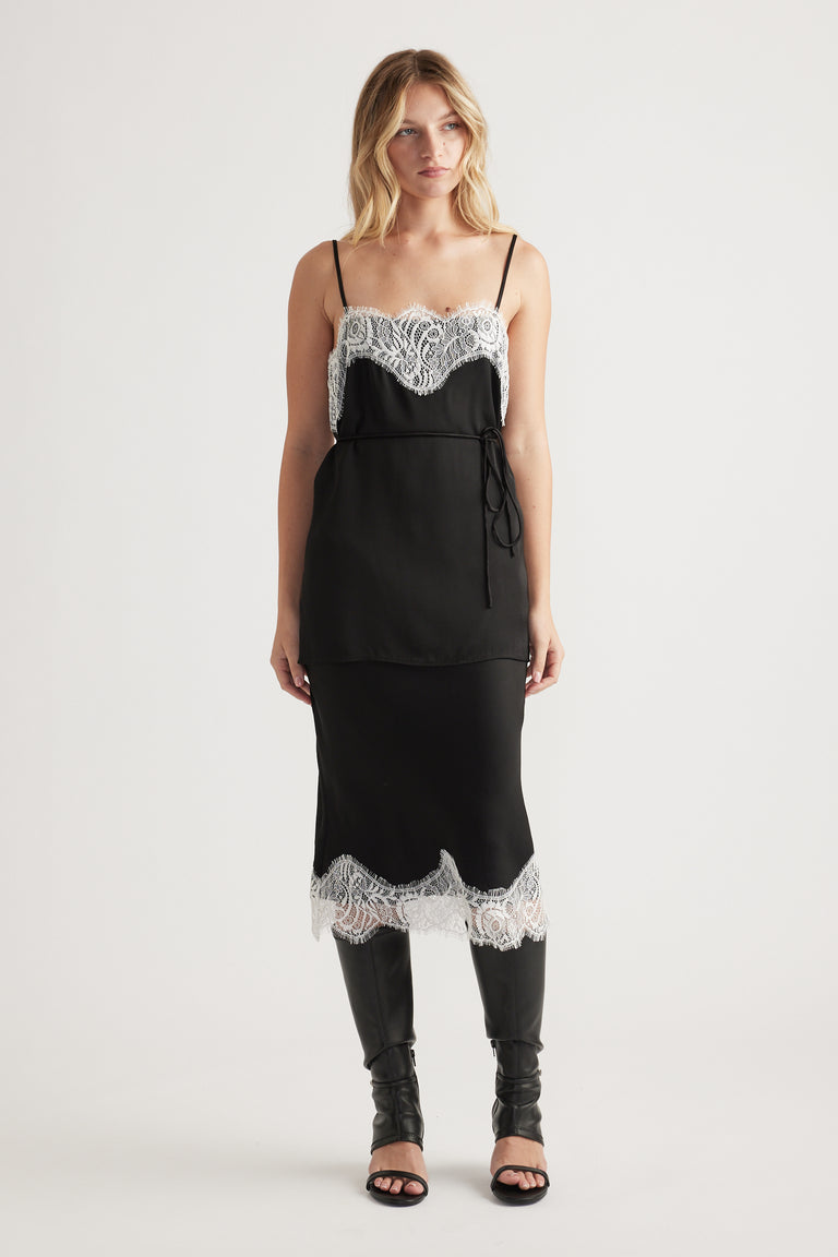 Woman wearing a black slip dress with white lace trim at neckline and hem, styled with tall black boots for an elegant look.