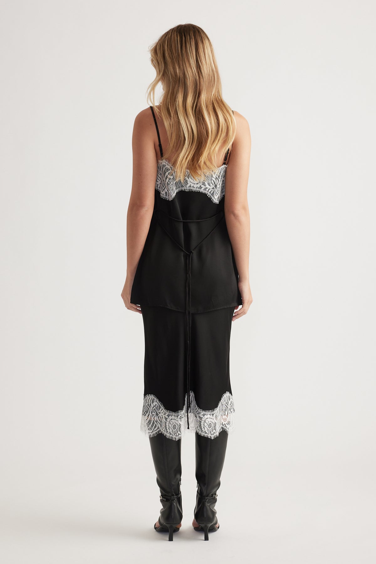Woman wearing a black slip dress with white lace trim on the back and hem, paired with black high boots.