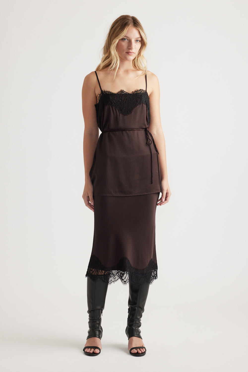 Woman wearing a dark lace-trimmed slip dress layered with a matching belted camisole and black knee-high boots.