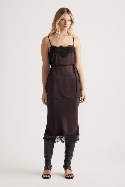 Woman wearing a dark lace-trimmed slip dress layered with a matching belted camisole and black knee-high boots.