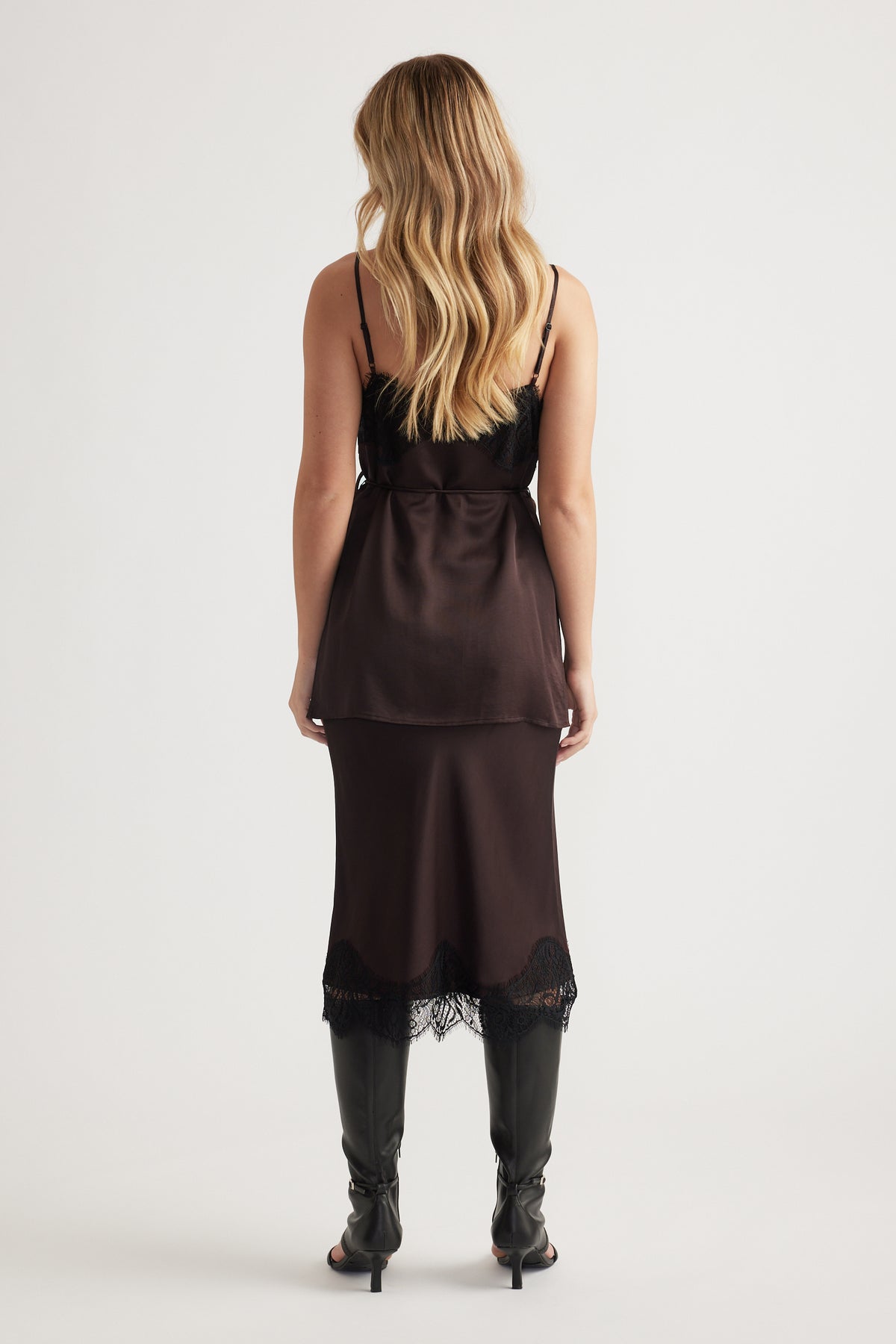 Woman wearing a dark lace-trimmed slip top paired with a matching skirt and black knee-high boots from behind.