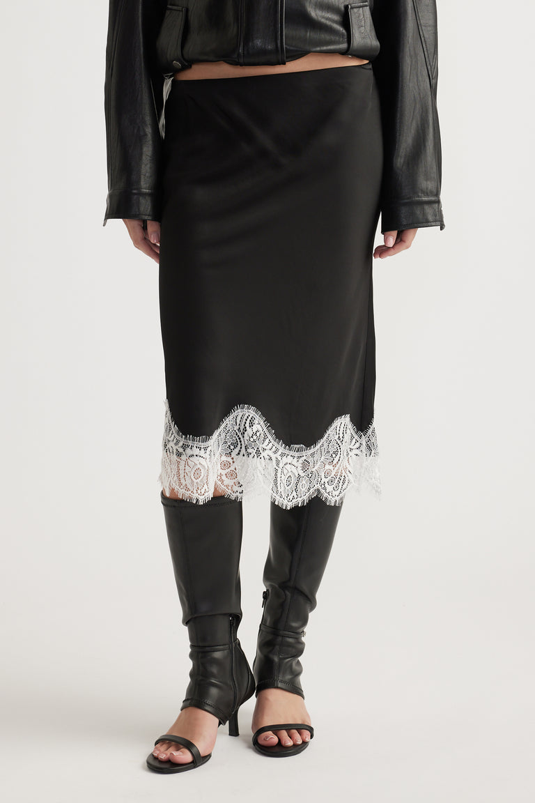 Black midi skirt with intricate white lace trim paired with knee-high black leather boots and a cropped jacket