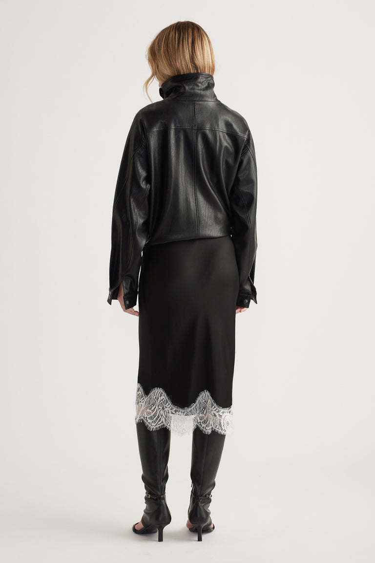 Woman seen from behind wearing a black leather jacket paired with a black skirt featuring white lace trim and heeled boots.