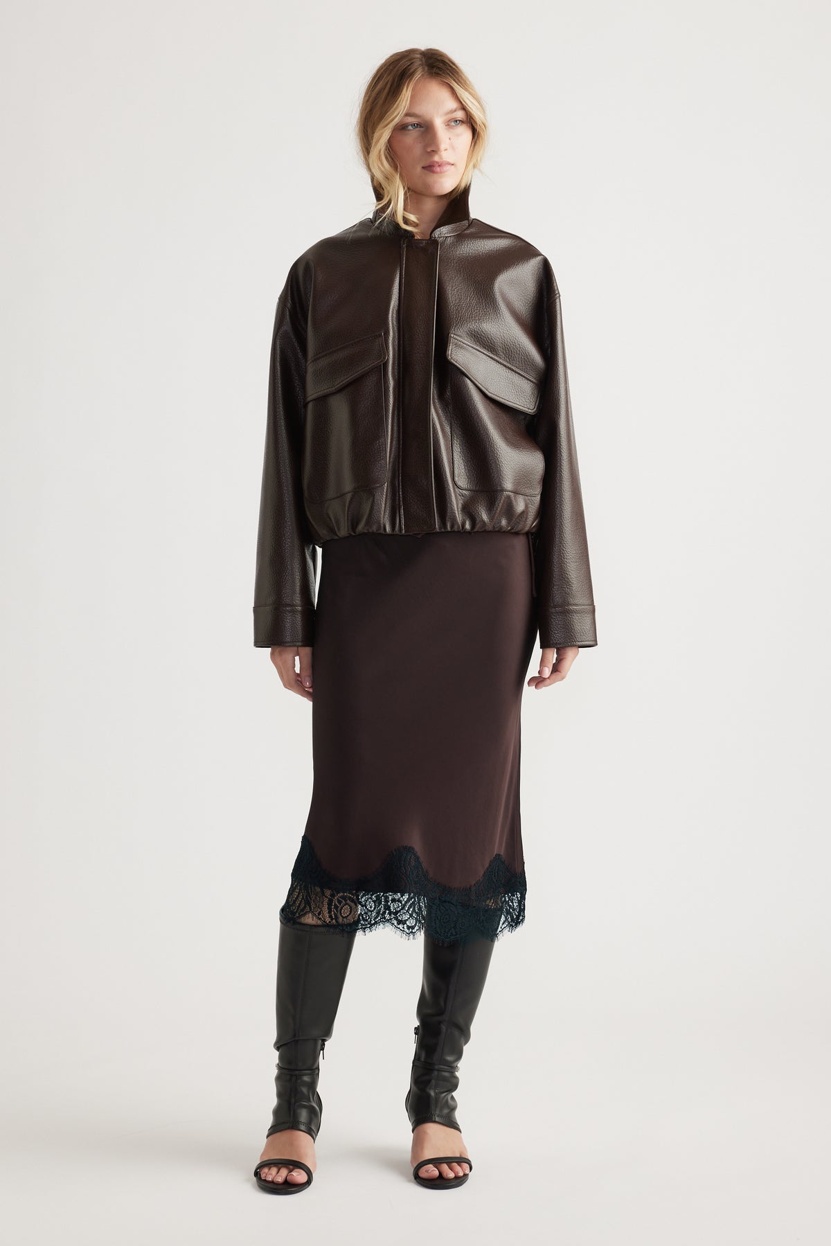 Woman wearing a dark brown cropped faux leather jacket over a midi skirt with lace trim and black knee-high boots.