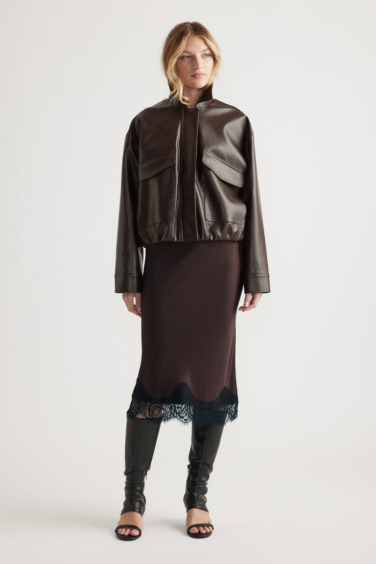 Woman wearing a dark brown cropped faux leather jacket over a midi skirt with lace trim and black knee-high boots.