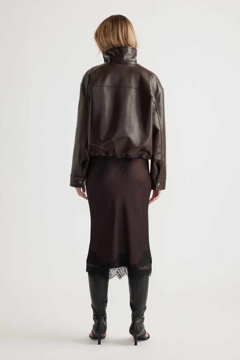 Woman seen from behind wearing a dark brown leather jacket paired with a midi skirt featuring lace trim and black heeled boots