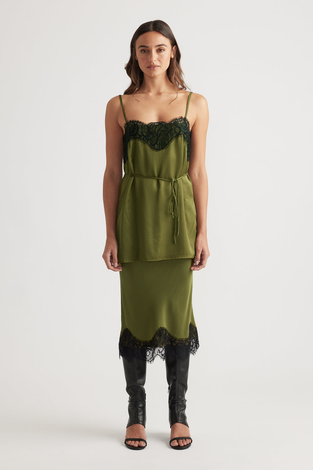 Front view of an olive green lace-trimmed slip dress layered with a matching tied waist top and black heeled boots
