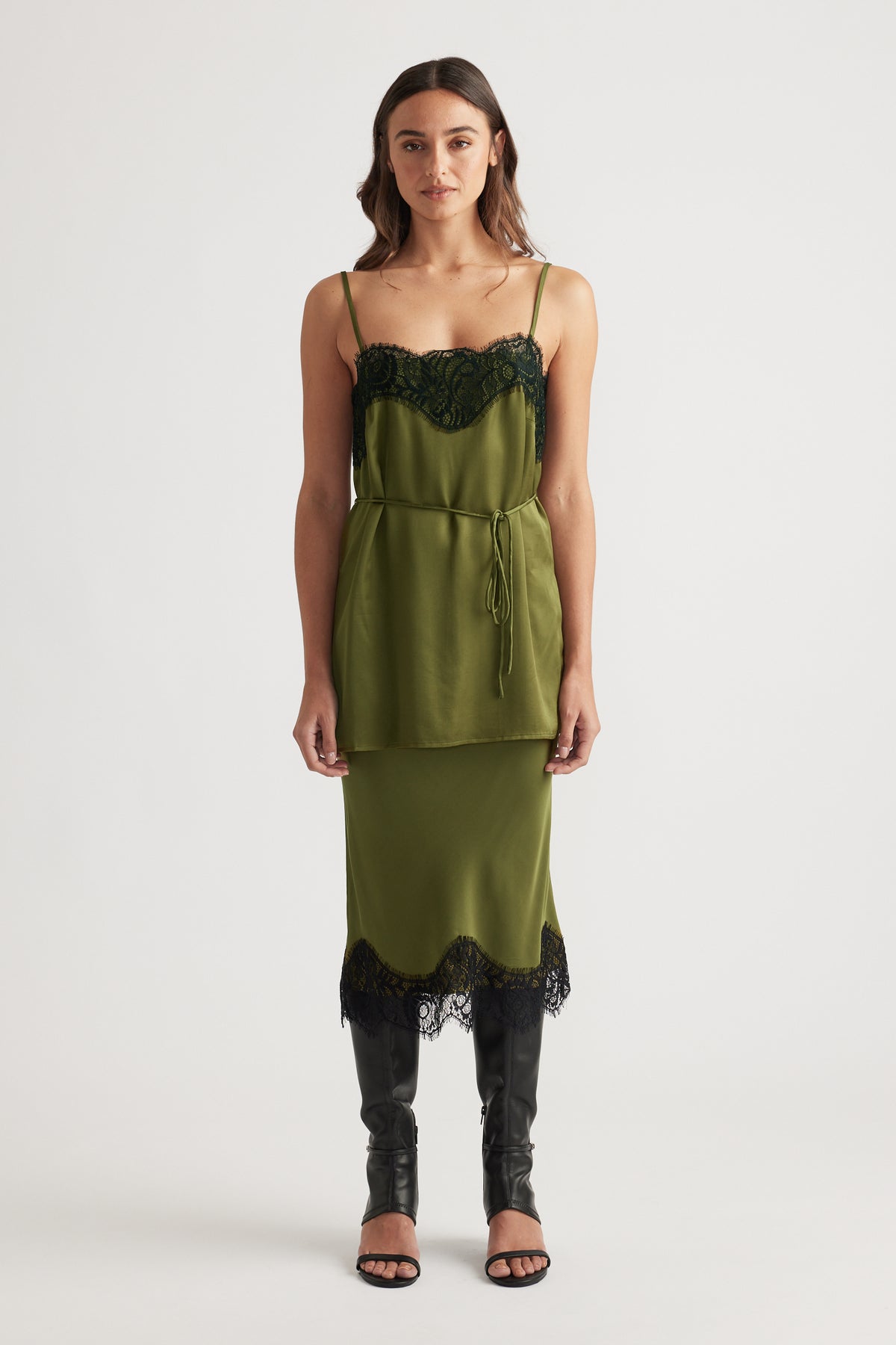 Front view of an olive green lace-trimmed slip dress layered with a matching tied waist top and black heeled boots