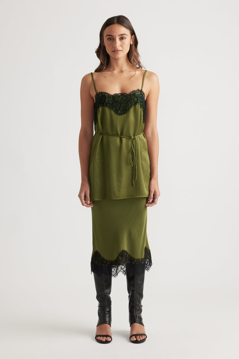 Front view of an olive green lace-trimmed slip dress layered with a matching tied waist top and black heeled boots