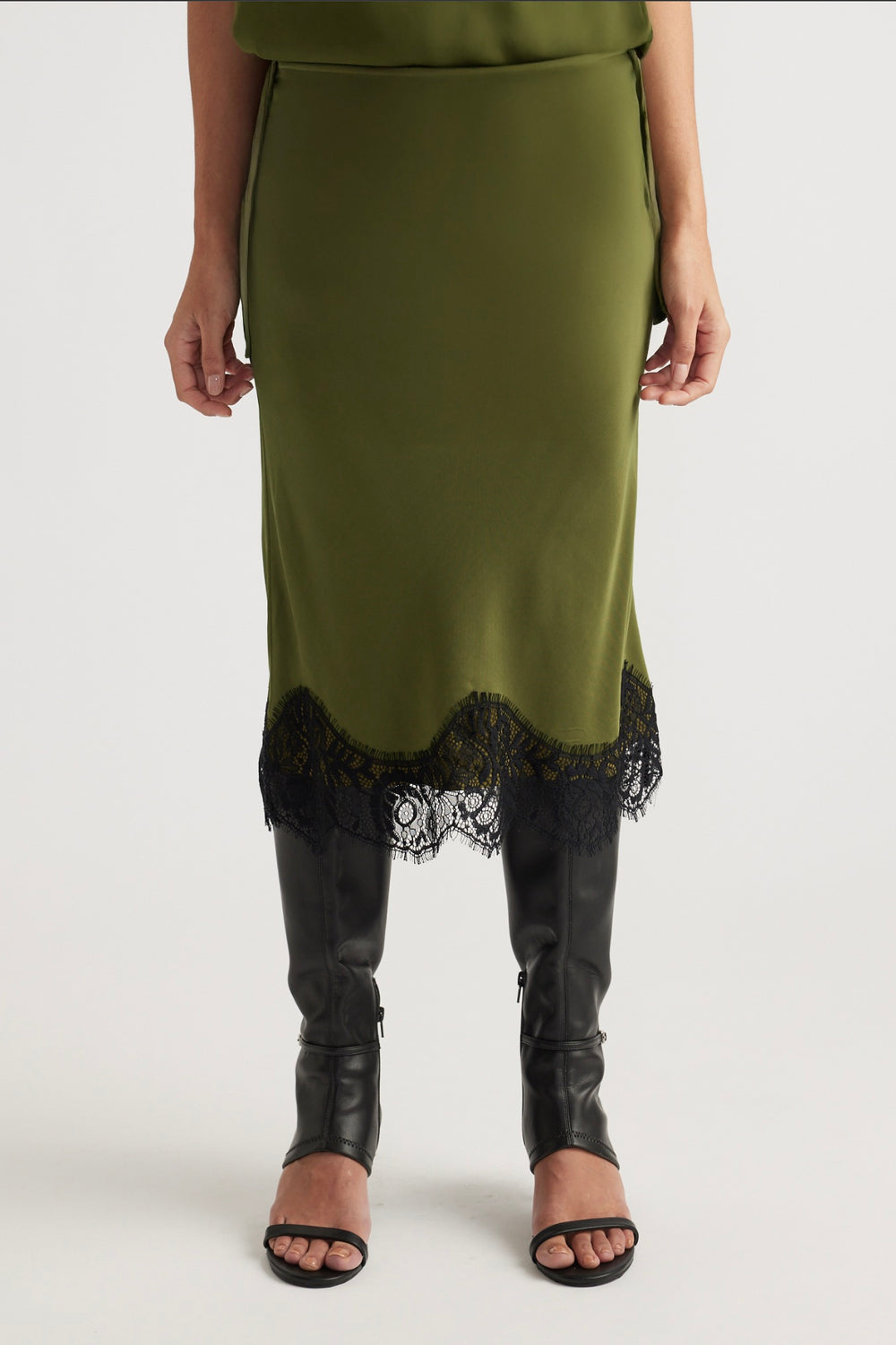 Front view of an olive green skirt with black lace trim paired with tall black leather heeled boots and sandals